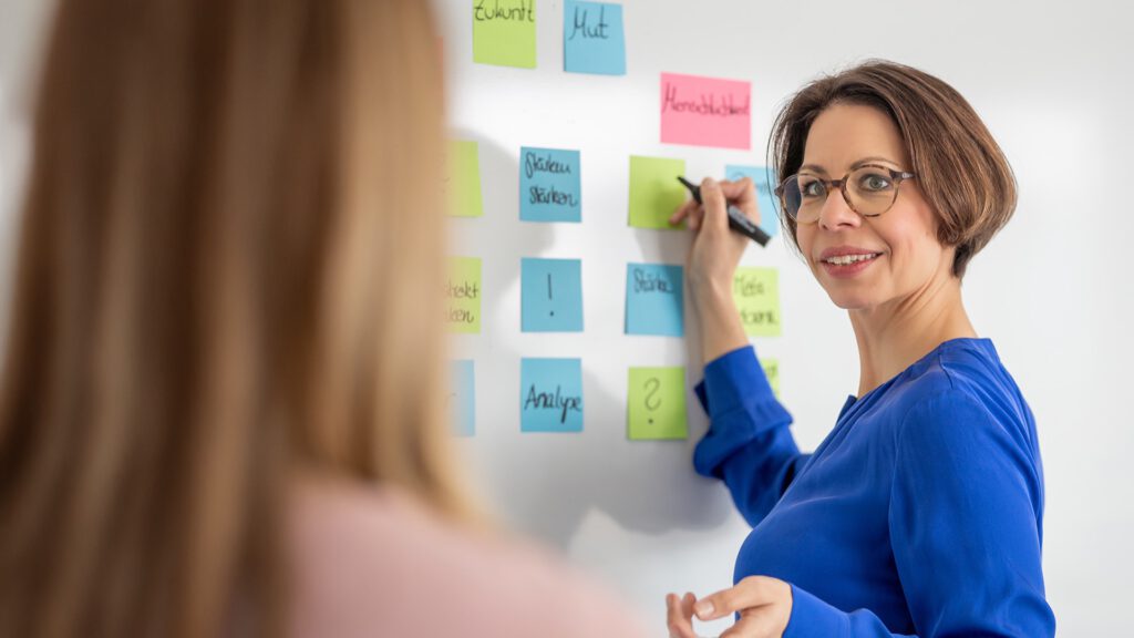 Elena Springub im blauen Kleid klebt bunte Notizzettel an ein Whiteboard und lächelt in die Kamera, im Vordergrund eine Kollegin.