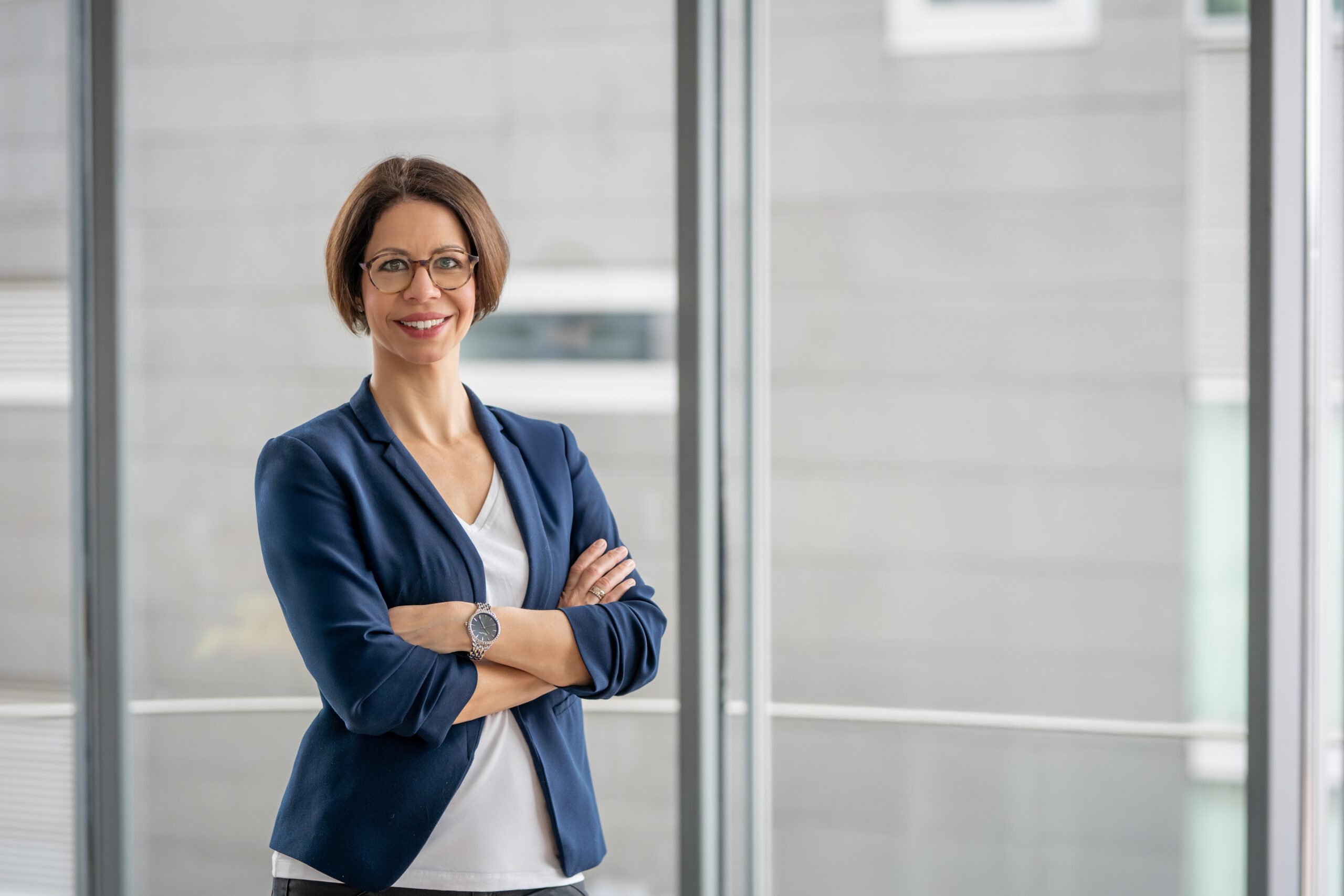 Elena Springub im blauen Blazer mit verschränkten Armen vor einer Fensterfront.