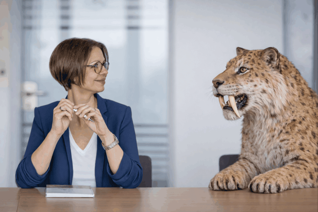 Elena Springub sitzt am Konferenztisch, neben ihr liegt ein Säbelzahntiger auf dem Tisch, Fotomontage.