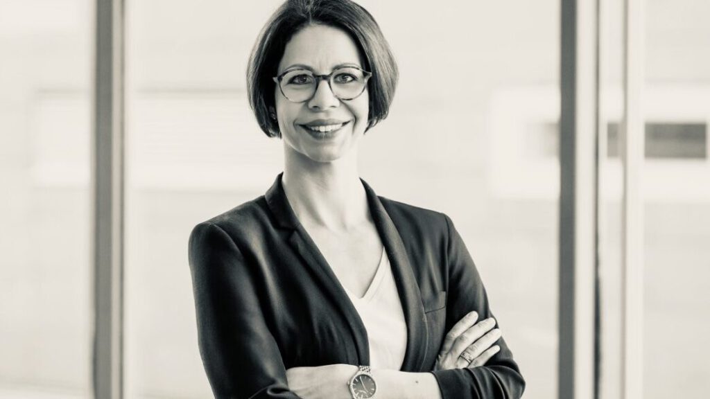 Elena Springub im dunklen Blazer mit verschränkten Armen, Schwarzweißportrait vor einer Fensterfront.